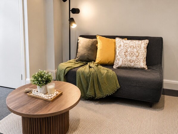 Un salón con un sofá gris oscuro, una mesa de centro redonda de madera y una manta verde.