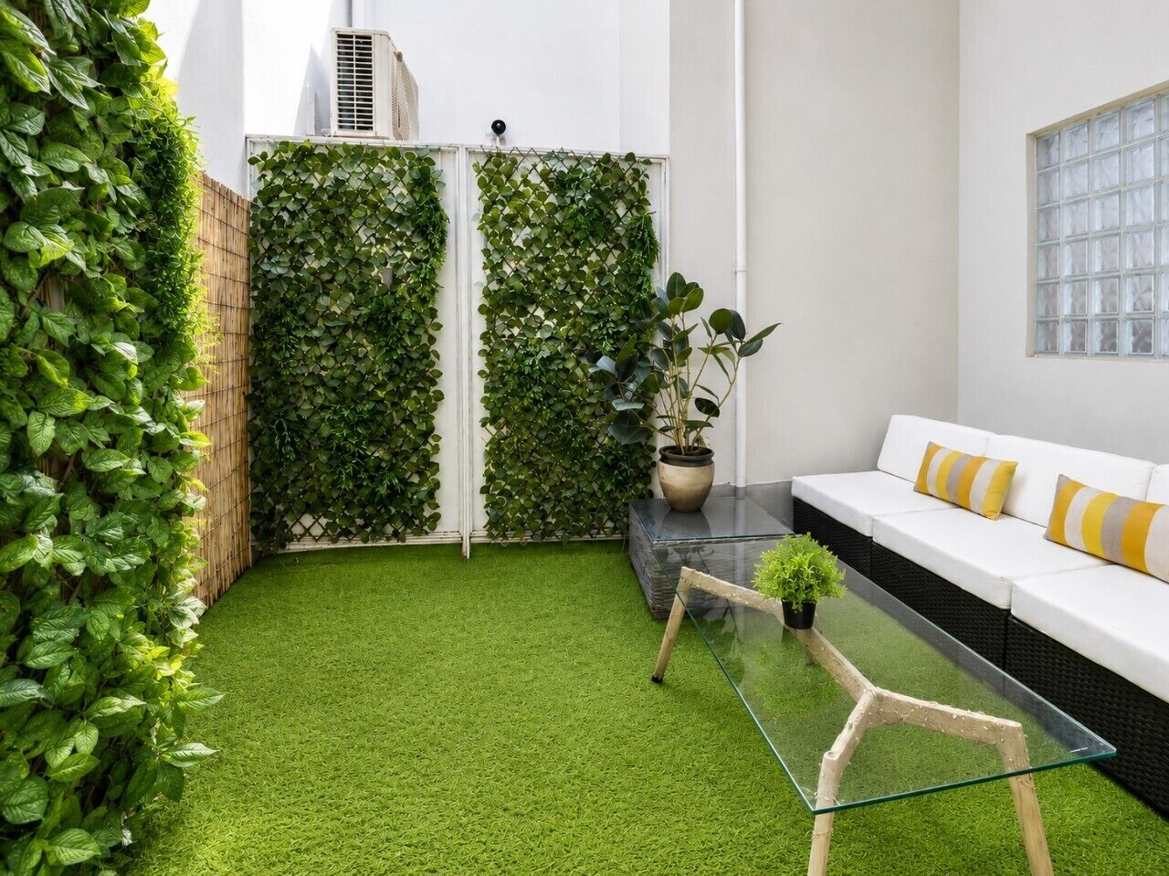 A modern outdoor seating area with a glass table, a white sofa with yellow cushions, and a green artificial grass floor.