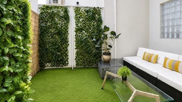 A modern outdoor seating area with a glass table, a white sofa with yellow cushions, and a green artificial grass floor.