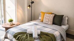 A bedroom with a bed, pillows, a bedside table with a plant, a floor lamp, and a wall-mounted artwork.