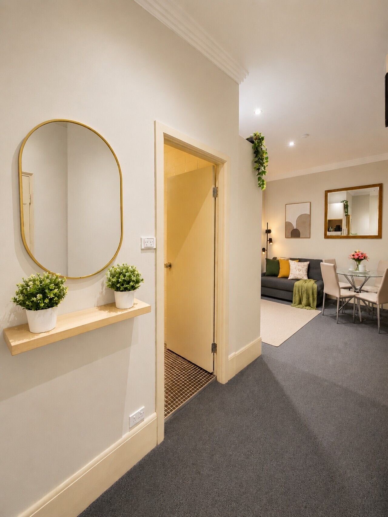 A hallway leading to a living area with a sofa, a dining table with chairs, a round mirror, and two potted plants on a shelf.