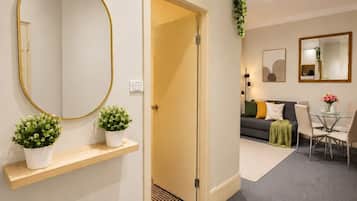 A hallway leading to a living area with a sofa, a dining table with chairs, a round mirror, and two potted plants on a shelf.