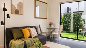 A living room with a sofa, coffee table, and a glass table with chairs. There is a large window with a view of a garden.