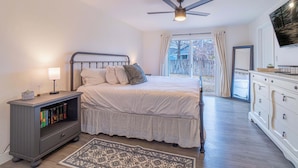 A bedroom with a bed, bedside table, bookshelves, a ceiling fan, and a large window.
