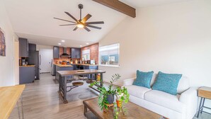 A modern kitchen with a dining area, a white sofa with blue cushions, and a wooden coffee table with plants.