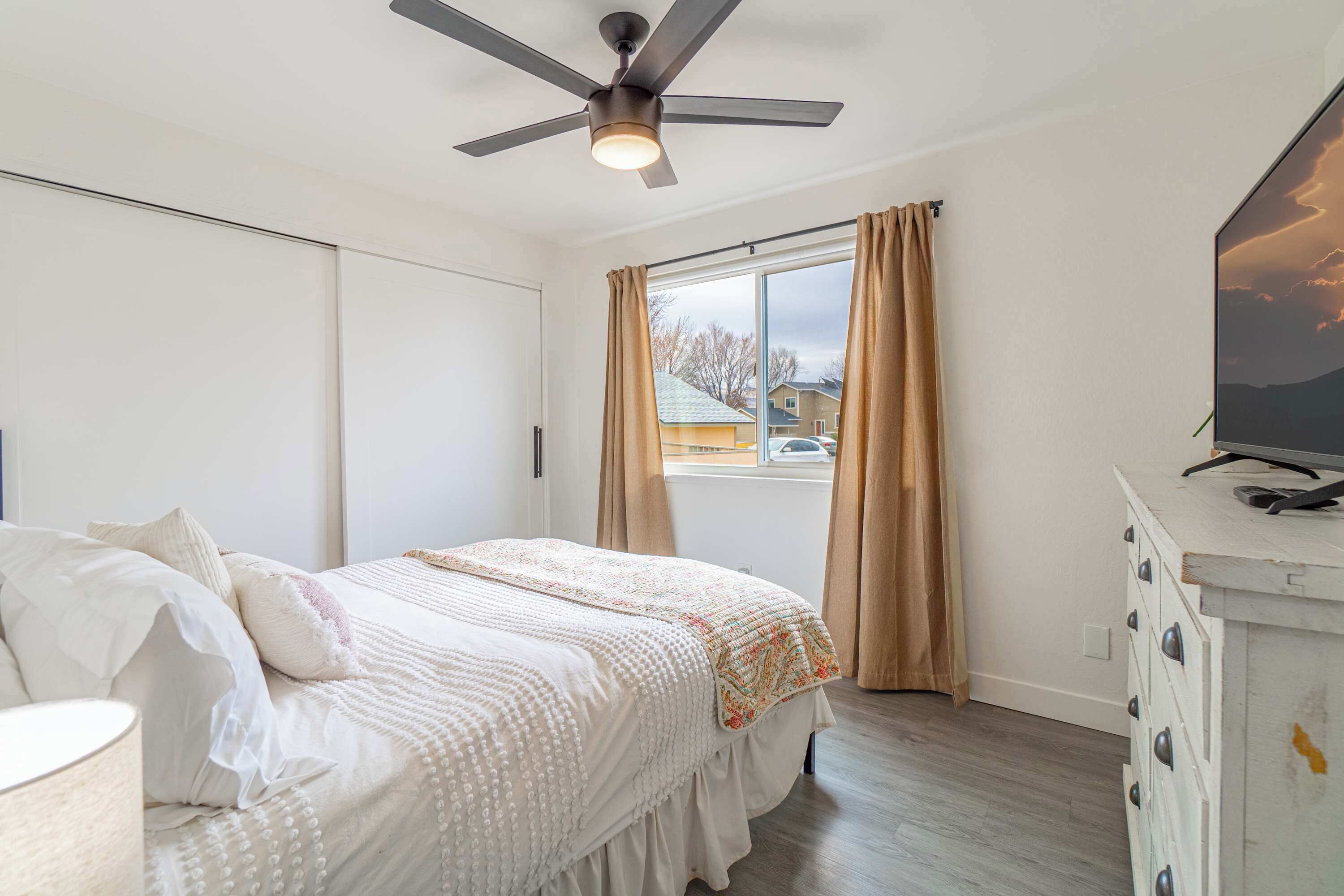A bedroom with a bed, a ceiling fan, a dresser, and a window with curtains.