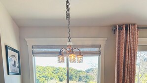 Ein Esszimmer mit Holztisch, vier Stühlen, einer Chandelierleuchtre und einem Fenster mit Blick ins Grüne.
