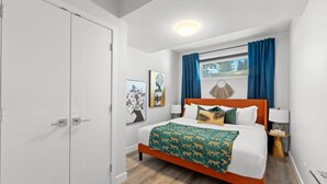 A bedroom with a bed, a nightstand, a window with curtains, a door, and a wall with a picture. - The Den | Centrally Located | King Bed | WiFi! (Edmonton)