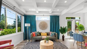 A spacious living room with a large window, a sofa with colorful cushions, a round coffee table, and a dining area with chairs and a table. - Jungle House | AirCon | Sleeps 8 | Near WEM & DT! (Edmonton)