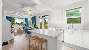 A modern kitchen with white cabinetry, a large island with stools, and a view of the outdoors. - Jungle House | AirCon | Sleeps 8 | Near WEM & DT! (Edmonton)