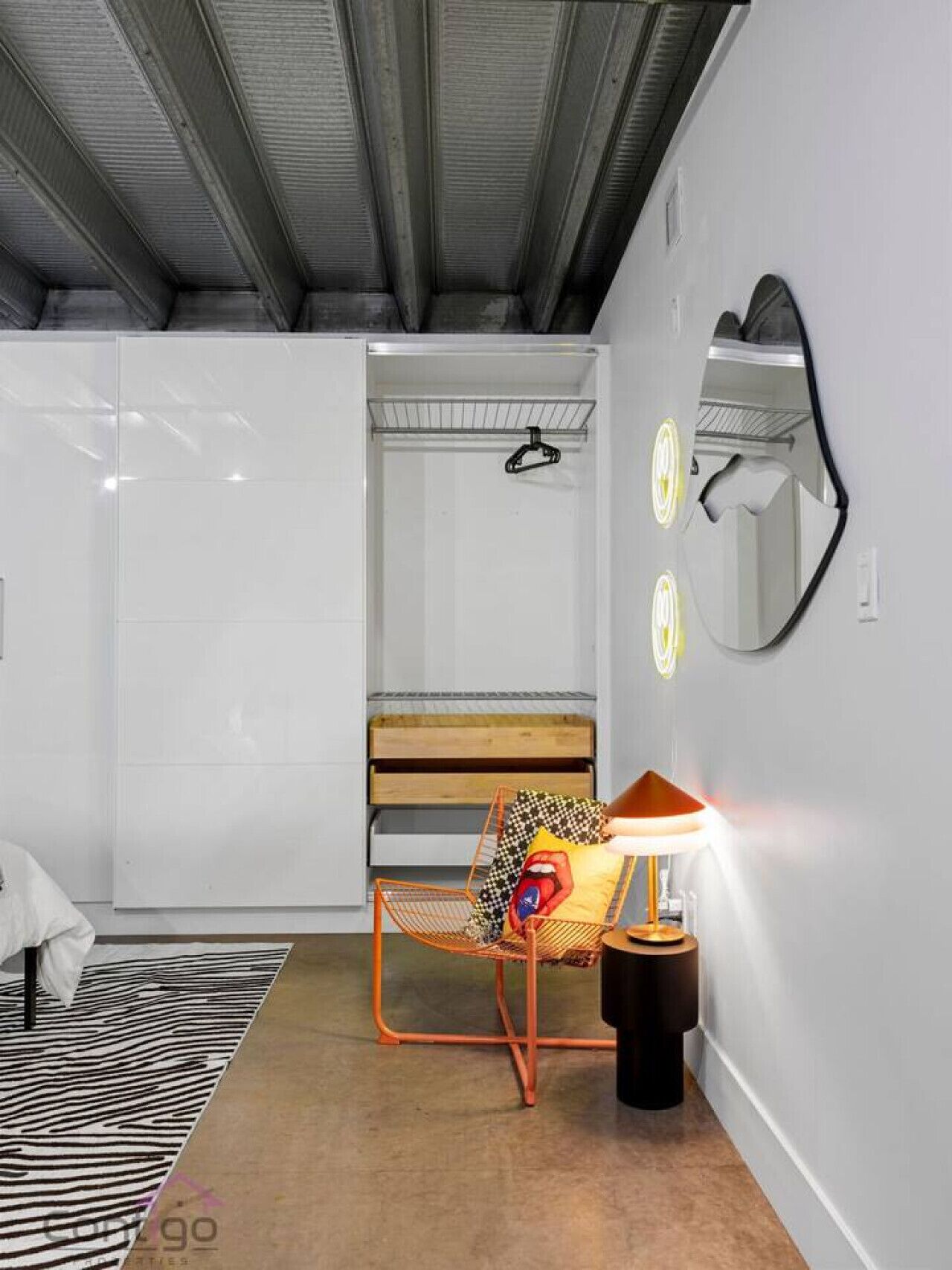 A modern hotel room with a metal-framed chair, a small table with a lamp, and a large mirror on the wall.