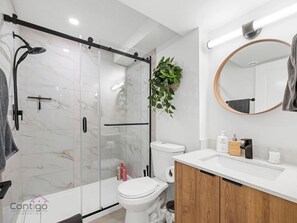 A modern bathroom with a glass shower enclosure, a toilet, a sink with a wooden cabinet, and a mirror with a towel hanging on it. - The Suite Escape | 2 BD | Near WEM & DT | Sleeps 5 (Edmonton)