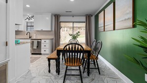Een eetgedeelte met een houten tafel en stoelen, uitzicht op de omgeving door een raam, en een groene wand met afbeeldingen in lijsten.