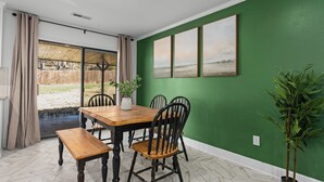 Een eethoek met een houten tafel en stoelen, een bank, een potplant en een groene wand met drie ingelijste foto's.