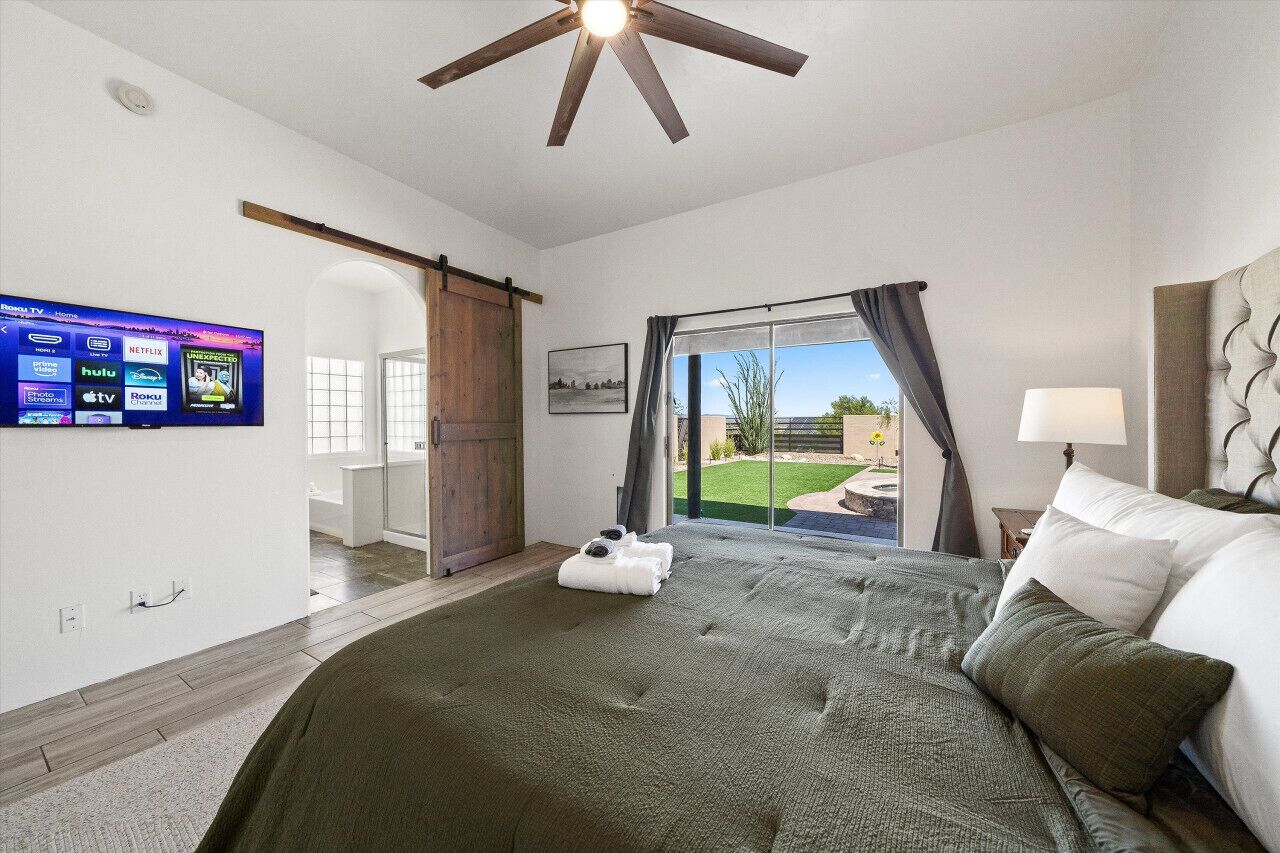 A bedroom with a large bed, a ceiling fan, a TV mounted on the wall, and a sliding glass door leading to a patio.