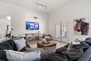 A modern living room with a large flat-screen TV, a comfortable sofa, a coffee table, and a horse painting on the wall. - 4BR Home w/ Mountain Views, Pool, Spa, Firepit & Gazebo (Tucson)