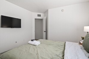 A hotel room with a bed, a flat-screen TV mounted on the wall, a bedside table with a lamp, and a door leading to another room.