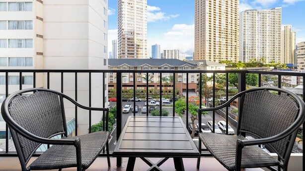A balcony with two wicker chairs and a table, overlooking a parking area and tall buildings. - NEW! Stylish and Cozy Studio near Waikiki Beach! (Honolulu)