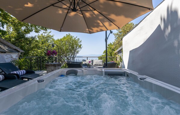 A hot tub with a view of the sea, surrounded by lounge chairs and an umbrella.