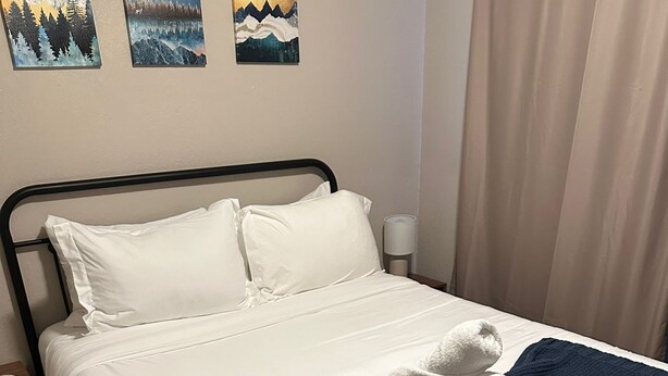 A neatly made bed with white linens and a dark blue blanket, a bedside lamp, and two landscape paintings on the wall. - The Denali Nook in UMED District & University prk! (Anchorage)