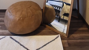 A room with a wooden floor, a brown leather ottoman, a white rug, and a full-length mirror reflecting a bed and curtains. - The Denali Nook in UMED District & University prk! (Anchorage)