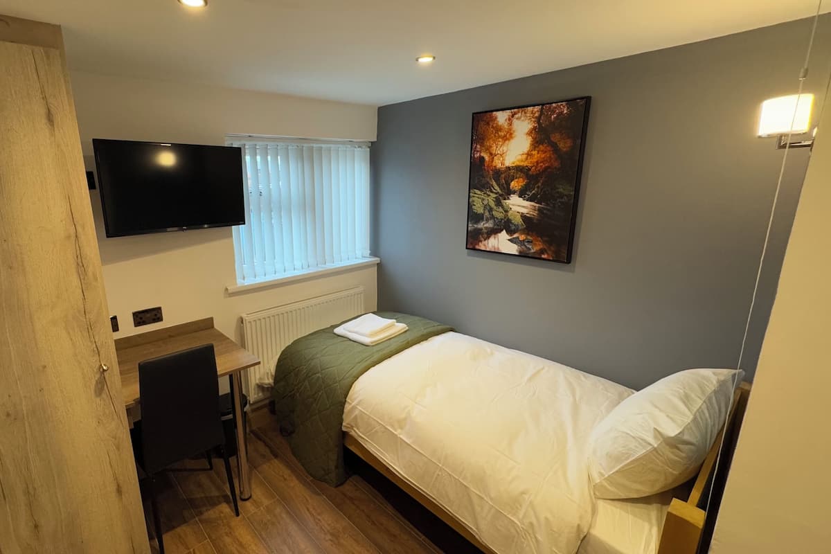 A compact bedroom with a bed, a desk, a chair, a TV mounted on the wall, and a window with blinds.