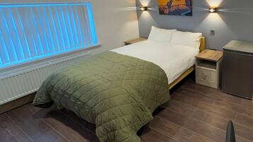A bedroom with a bed, a nightstand, a small refrigerator, a window with blinds, and a wall-mounted picture.