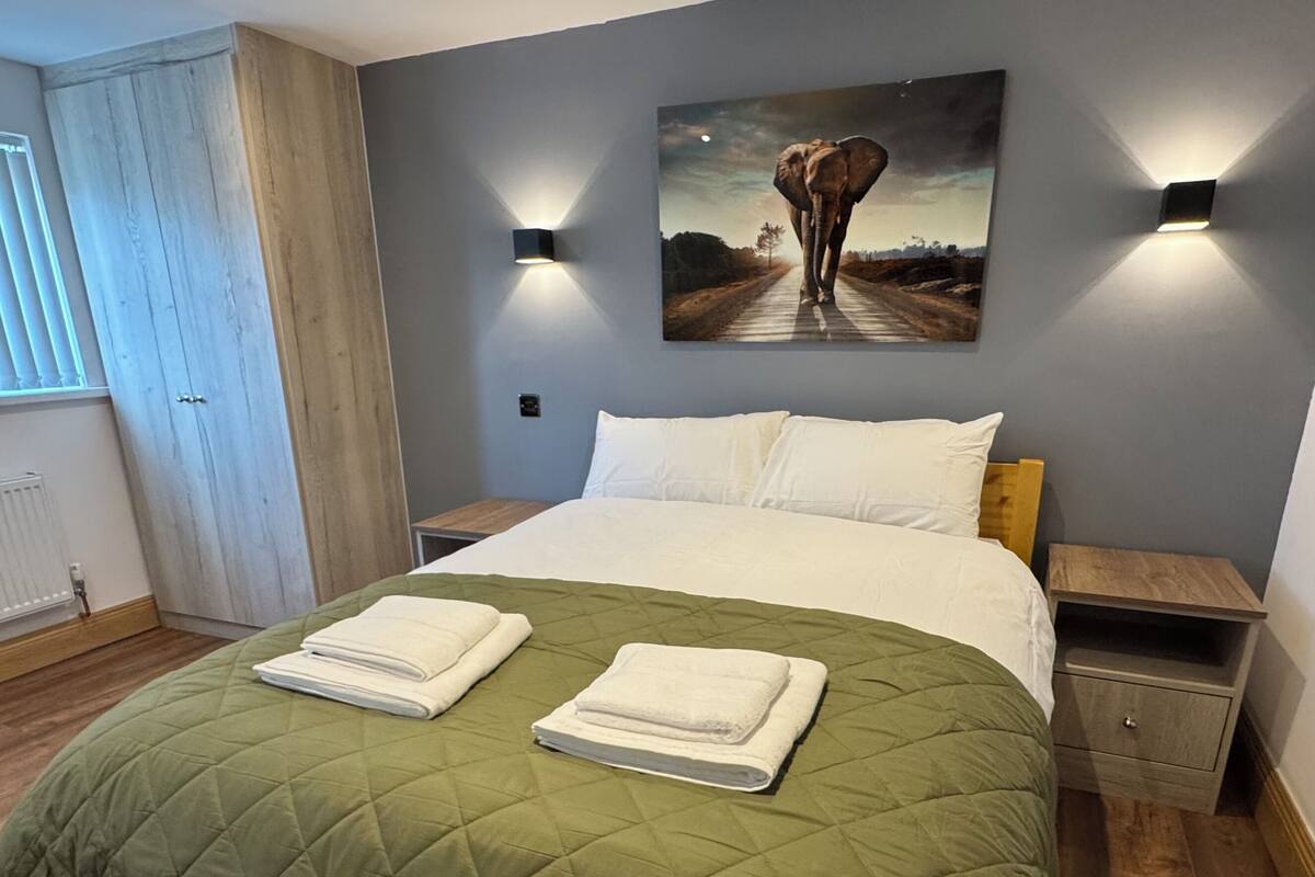 A neatly made bed with a green quilt, two bedside tables with lamps, a painting of an elephant on the wall, and a wooden floor.
