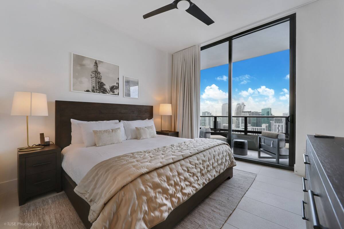 Ein modernes Hotelzimmer mit einem großen Bett, zwei Nachttischen mit Lampen, Blick auf die Stadt durch ein großes Fenster und einem Deckenventilator.
