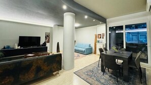 Un salon moderne doté d’un téléviseur à écran plat, d’un canapé de couleur foncée, d’un divan bleu clair, d’un coin repas avec une table et des chaises, et d’une grande fenêtre donnant sur un immeuble.
