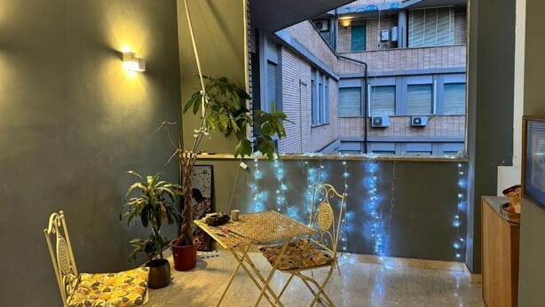 Un balcon avec une table et des chaises, des plantes en pot, des guirlandes lumineuses et une vue sur un immeuble.