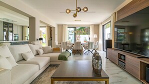 Un salon moderne avec un canapé d’angle, une table basse en bois, un coin repas avec une table et des chaises, un grand téléviseur à écran plat et une cuisine ouverte sur le salon par une porte.