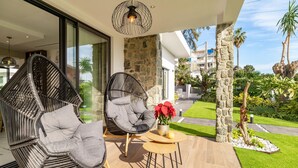 Un espace extérieur moderne, aménagé avec des chaises en osier et une table ronde, offrant une vue sur un jardin bien entretenu et un immeuble résidentiel.