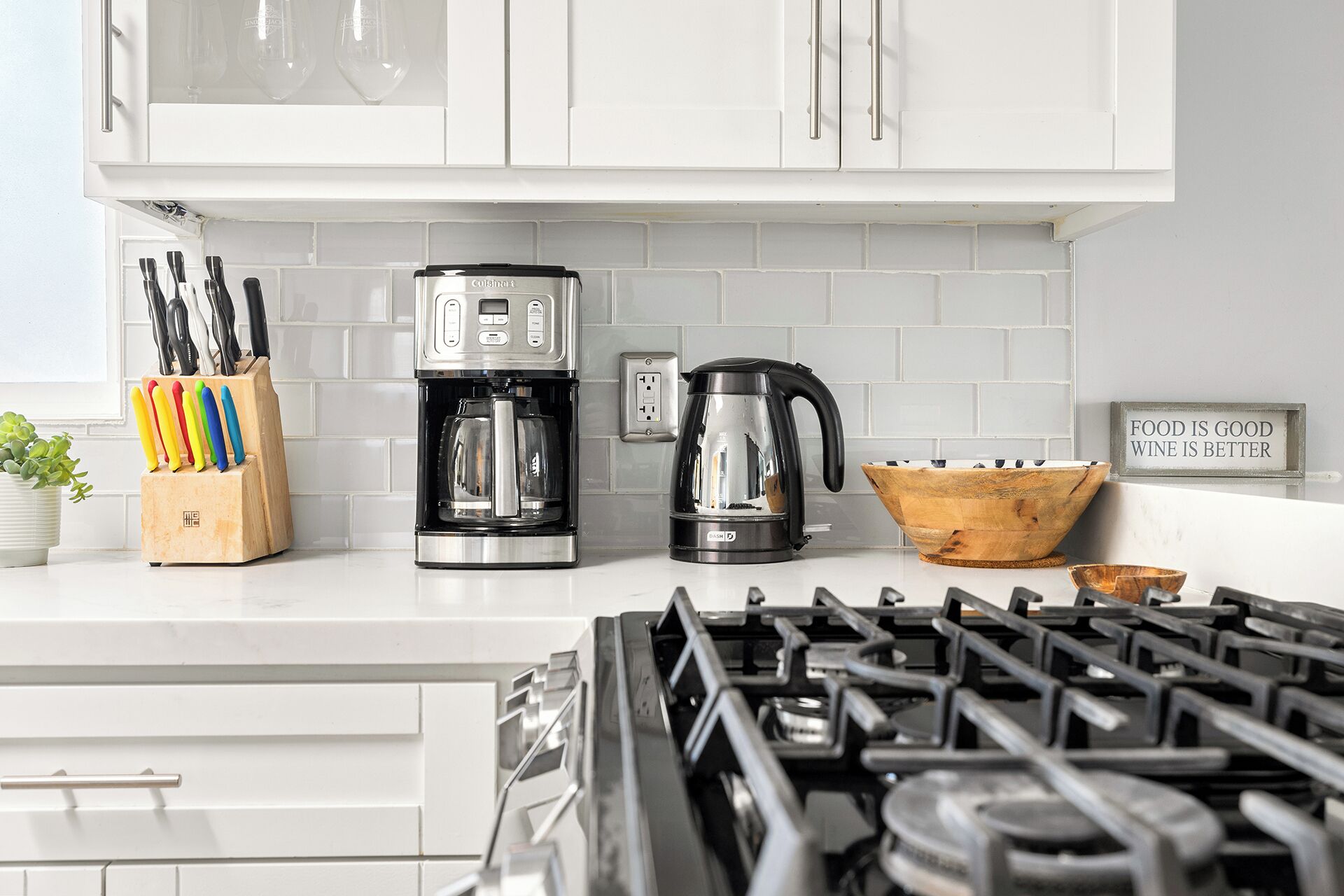 A kitchen with white cabinets, a coffee maker, a kettle, and a sign that says "FOOD IS GOOD WINE IS BETTER".
