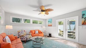 A spacious living room with a ceiling fan, orange sofas, a glass coffee table, and large windows offering a view of the outdoors. - Spacious and Short Drive to Beach (St. Petersburg)