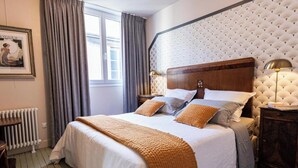 A neatly made bed with a headboard, a bedside table with a lamp, a radiator, and a window with curtains. - L' Appart 1925 - REIMS (REIMS)
