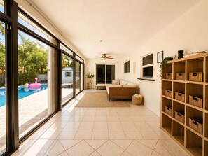 Een ruime woonkamer met een grote schuifpui naar een zwembad, een plafondventilator, een bank en een houten kastenwand.