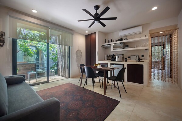A modern living room with a sofa, a dining area with chairs, a kitchen with shelves, and a ceiling fan.