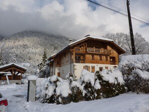 Una cabaña de madera con balcón, rodeada de árboles cubiertos de nieve y una montaña al fondo.