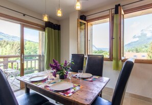 Una mesa de comedor dispuesta con platos y vasos, un jarrón con flores, y una vista de un paisaje montañoso a través de amplios ventanales.