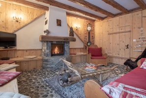 A cozy room with a stone fireplace, wooden beams, a television, and a stone floor. - Apartment 'Le Pele - 7 Pers' with Mountain View, Private Garden and Wi-Fi (Taninges)