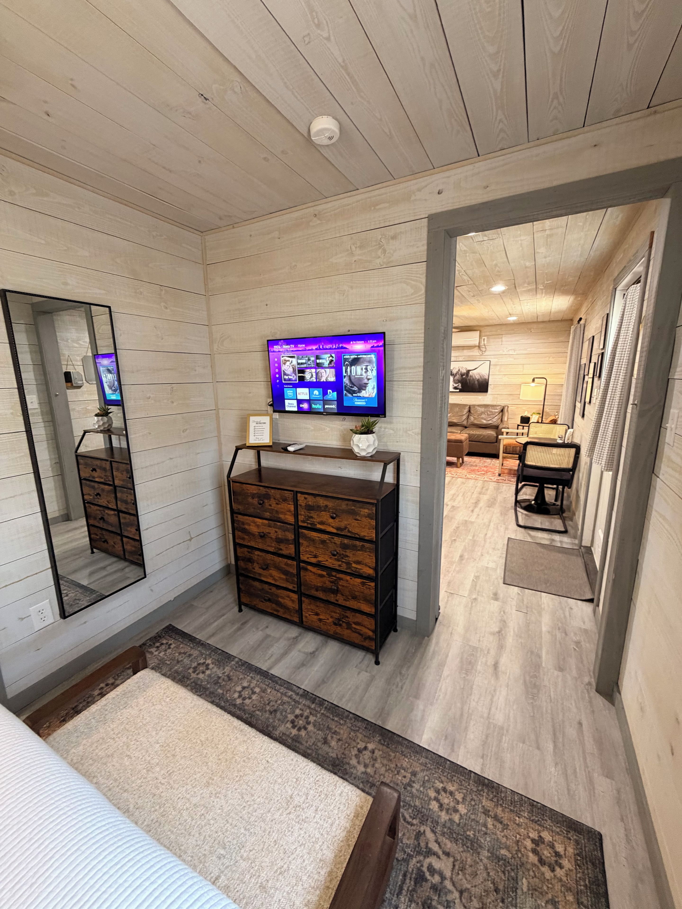 Een kamer met een houten plafond, een aan de muur bevestigde televisie, een dressoir, een spiegel en een deur naar een andere kamer.