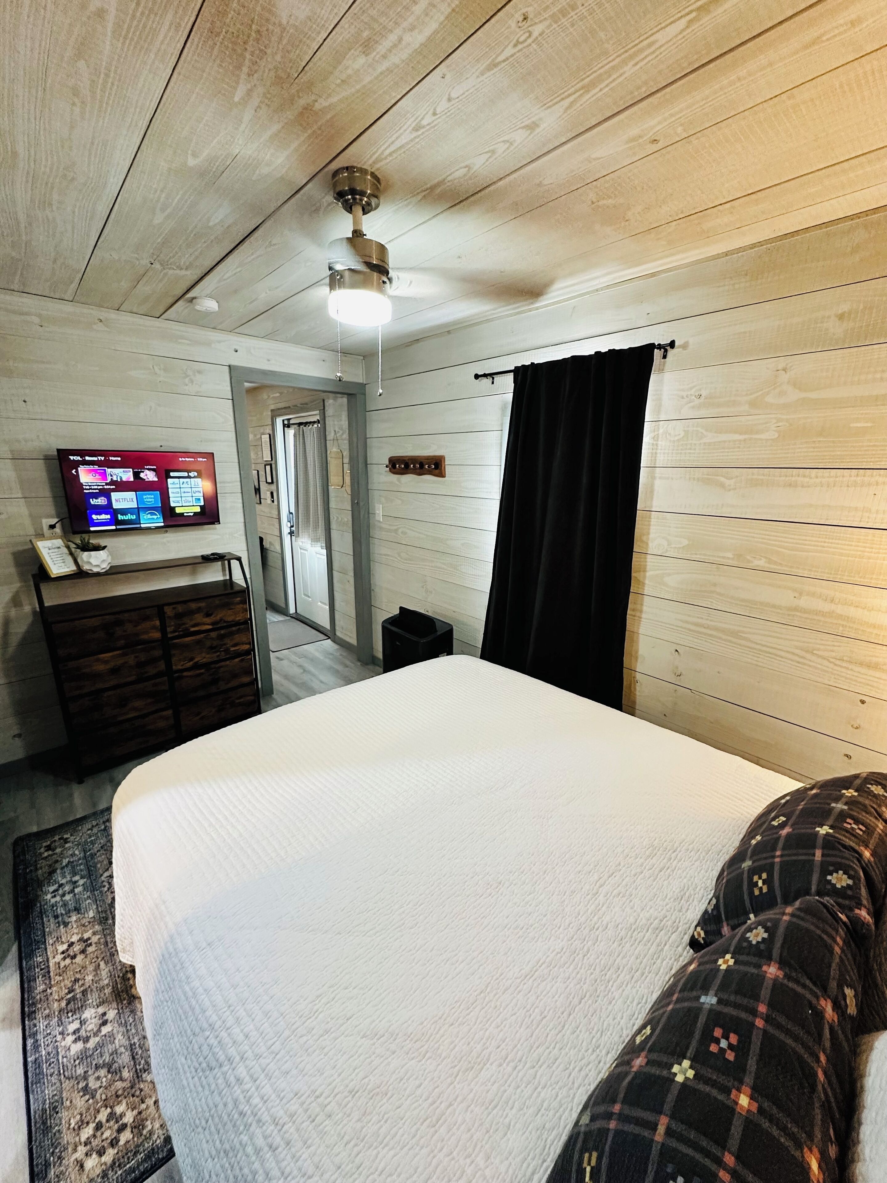 Un dormitorio con una cama, un televisor montado en la pared, techo de madera y una puerta.