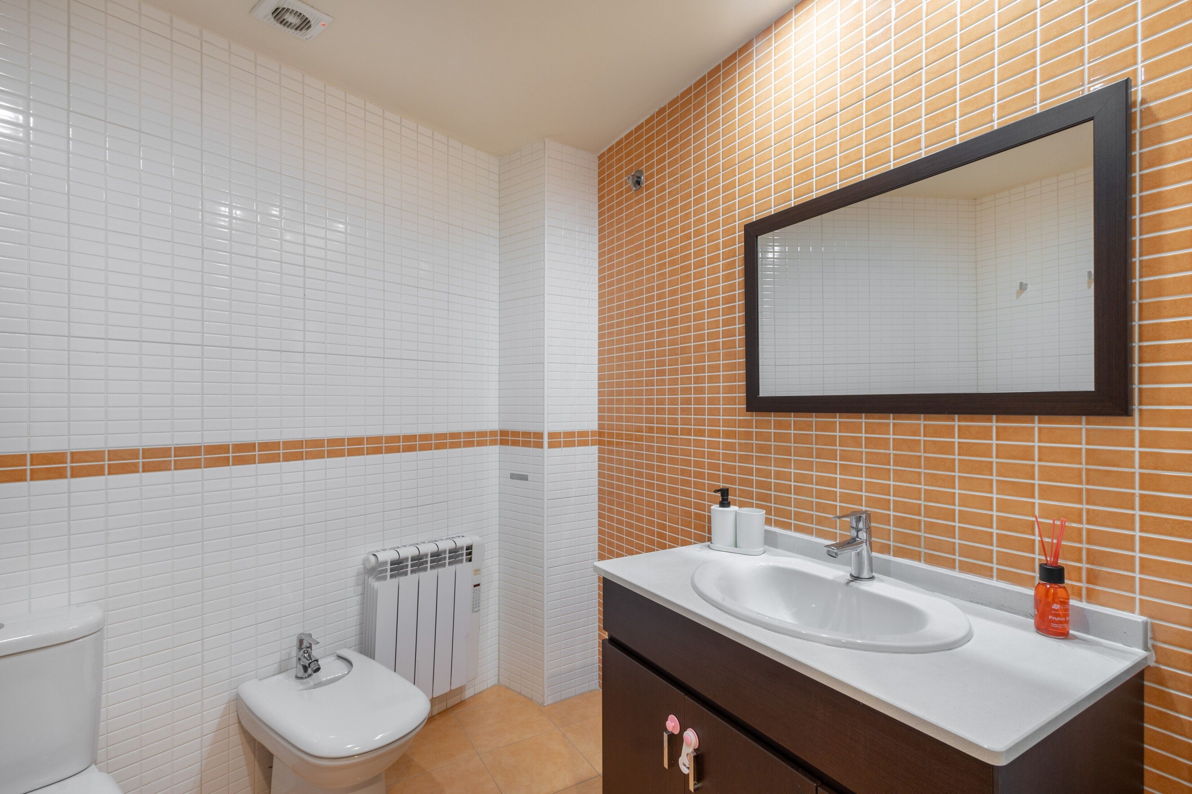 A bathroom with white tiles, a brown-tiled accent wall, a large mirror, a sink, a toilet, and a radiator.
