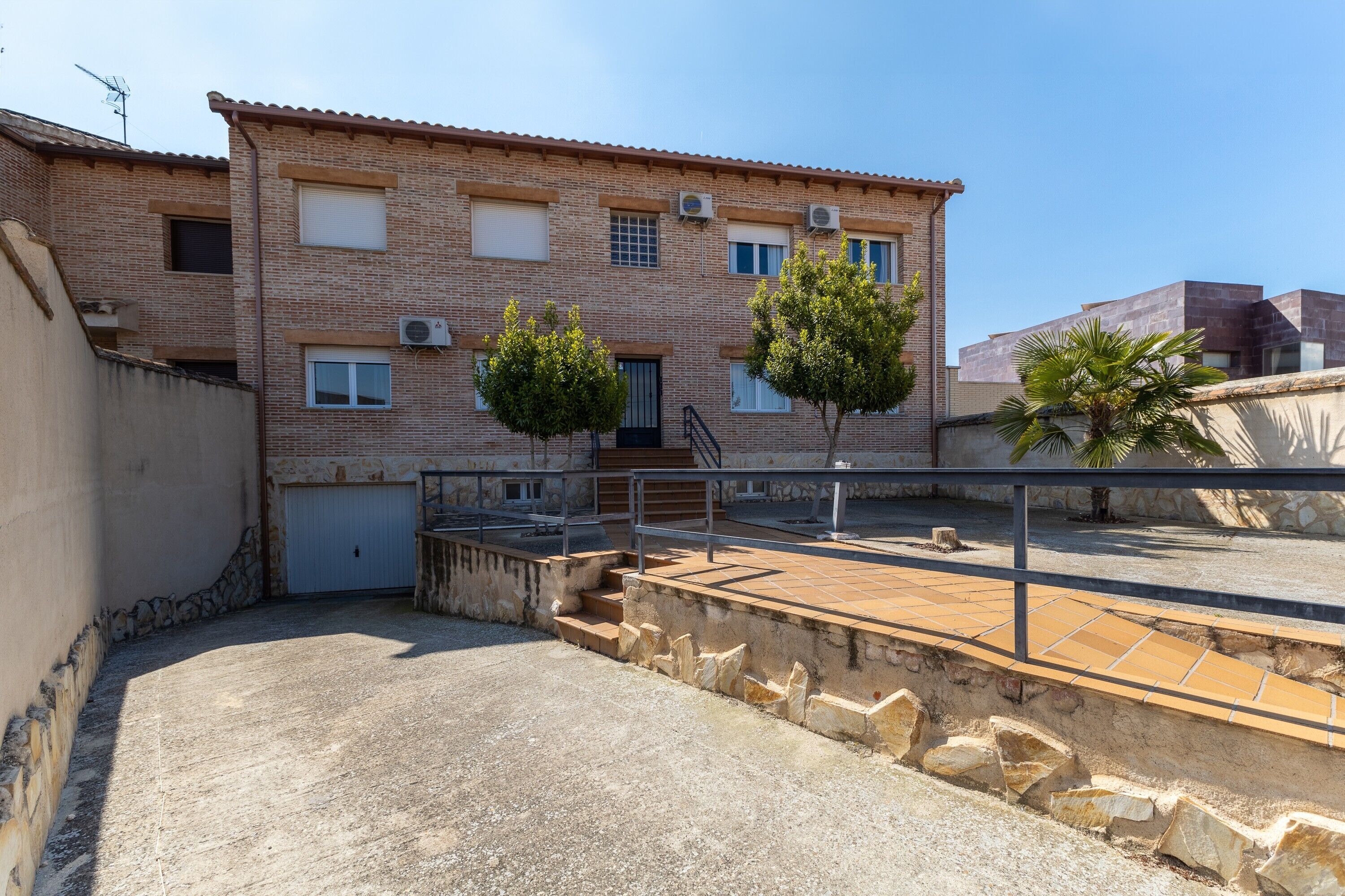 Un immeuble à plusieurs étages, doté d’un toit recouvert de tuiles, d’un garage et d’une cour pavée avec des marches menant à un balcon.
