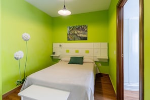 A bedroom with a bed, a bedside table, a hanging light fixture, a wall-mounted shelf, and a door leading to another room.