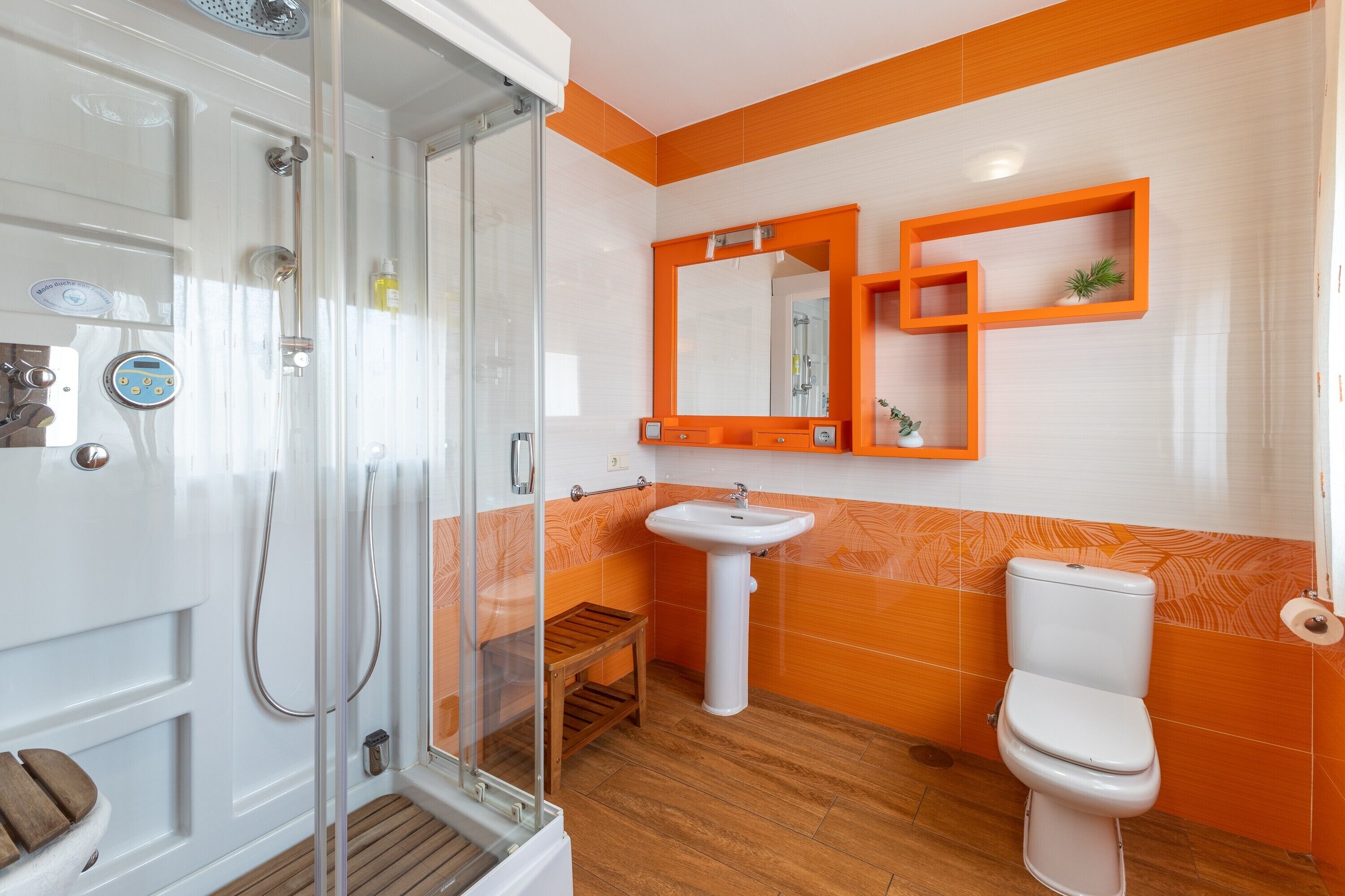 Un baño con cabina de ducha de vidrio, lavabo, inodoro y un pequeño taburete de madera.