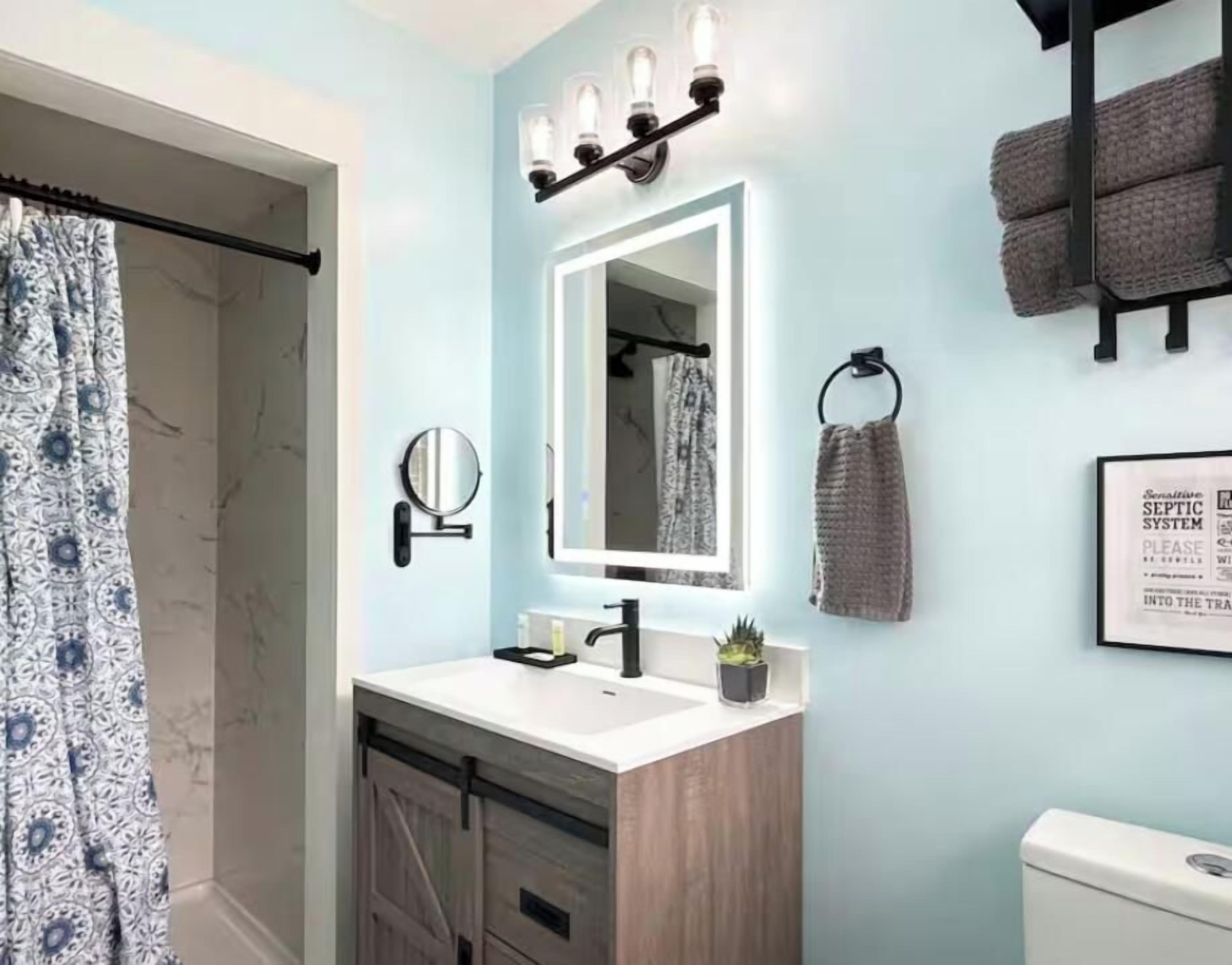 Un baño con pared azul claro, un lavabo blanco, un espejo con iluminación, un perchero para toallas y un letrero enmarcado.