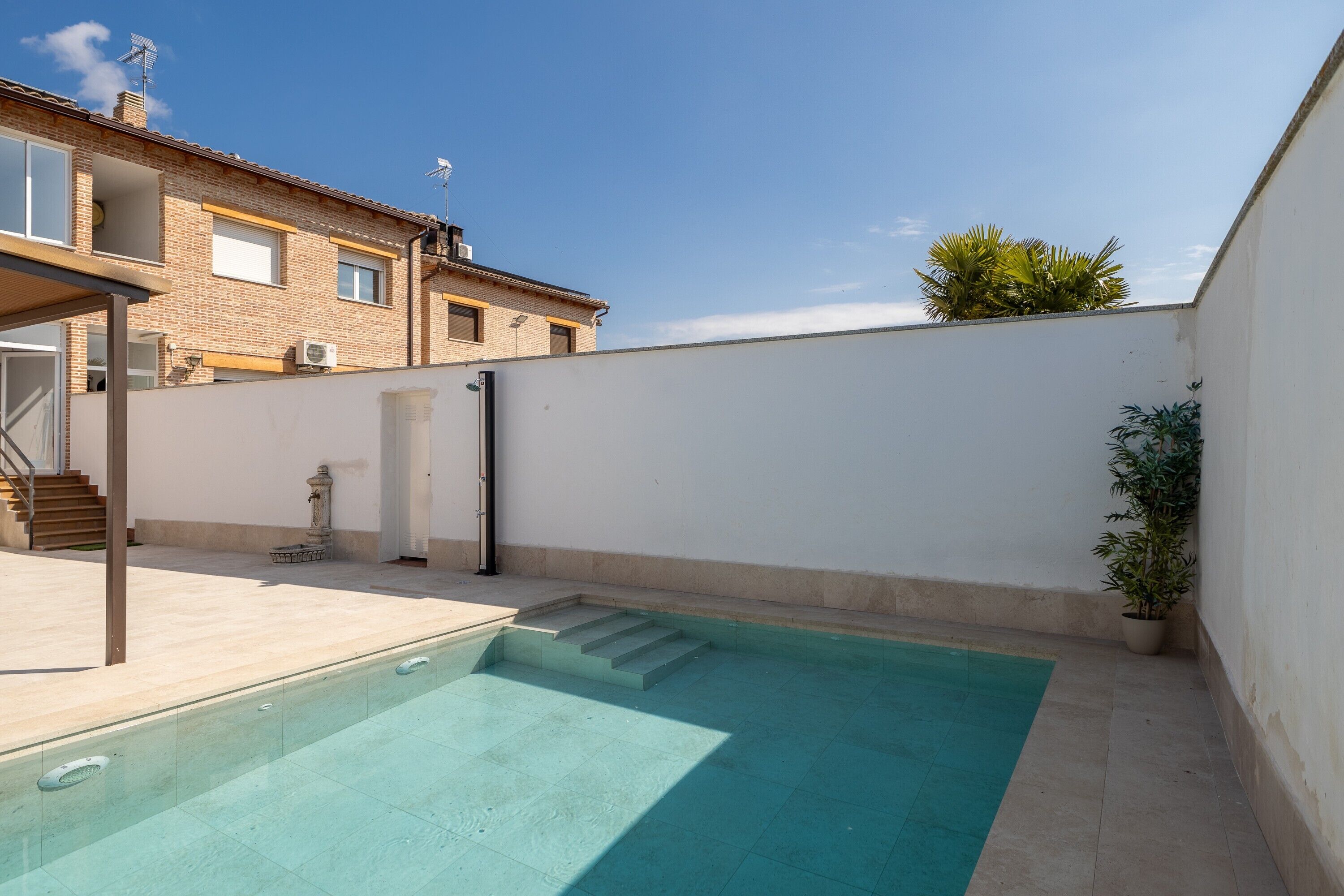 Une piscine avec des marches menant à l’intérieur, un mur blanc et, au fond, un bâtiment avec un toit recouvert de tuiles.