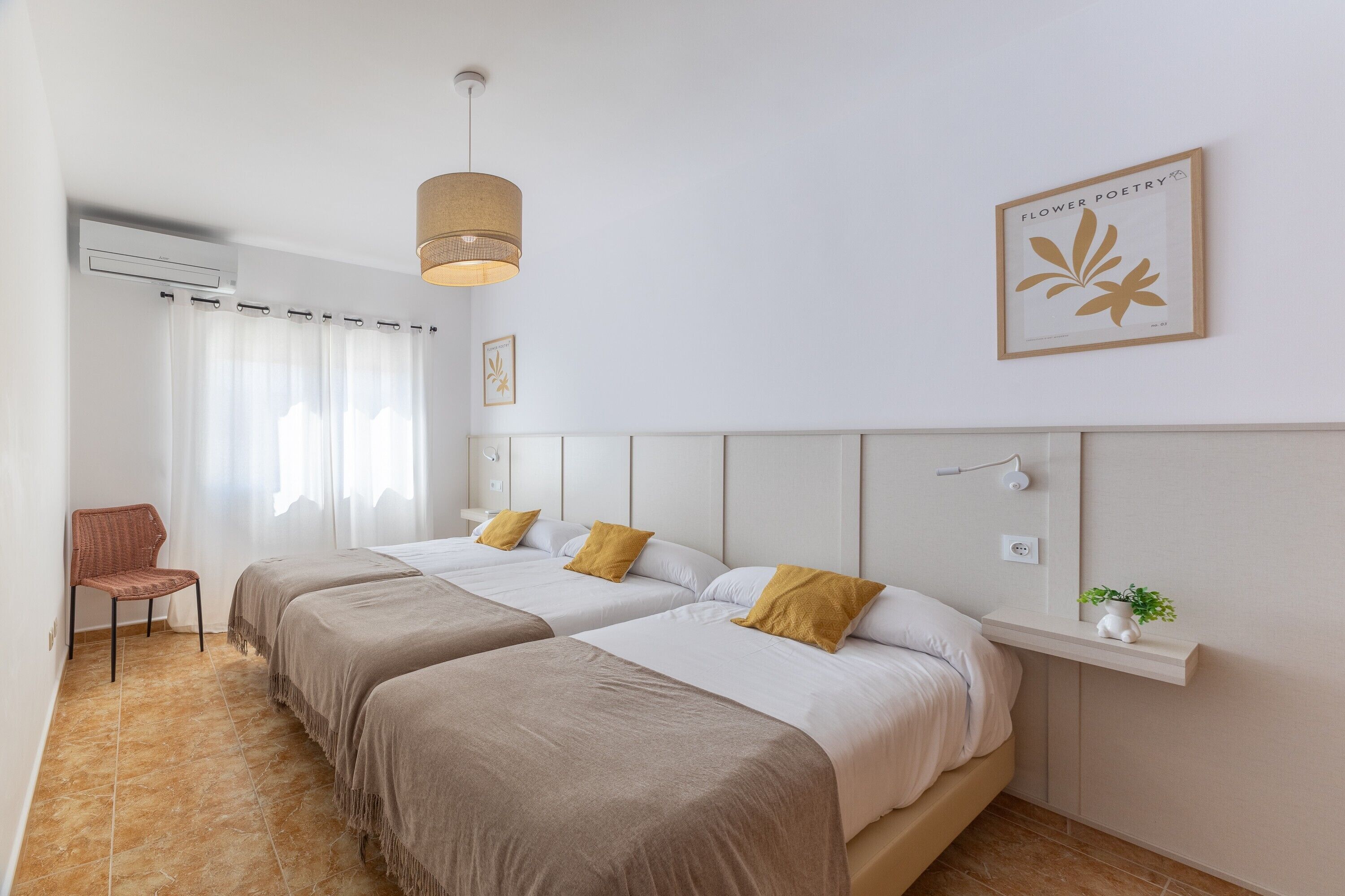 A hotel room with two beds, a chair, a hanging light fixture, a wall-mounted shelf with a plant, and a framed picture.
